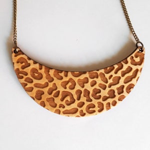 May include: A wooden necklace with a crescent-shaped pendant featuring a leopard print design. The pendant is made of light brown wood and has a brass chain.