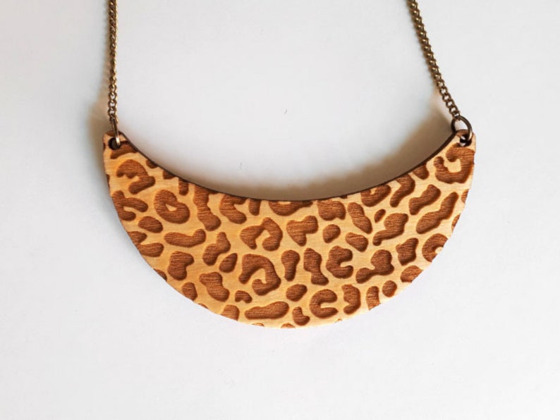 May include: A wooden necklace with a crescent-shaped pendant featuring a leopard print design. The pendant is made of light brown wood and has a brass chain.