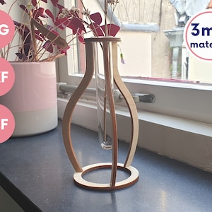 Op de afbeelding: Een houten vaas met een ronde basis en een cilindrische opening voor een reageerbuis. De vaas is gemaakt van 3 mm materiaal en wordt getoond op een zwart oppervlak met een plant in een roze pot op de achtergrond.