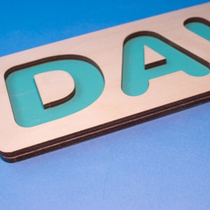 Op de afbeelding: Houten naam puzzel met de letters "DAVID" in turquoise in een natuurlijke houten lijst. De puzzel is ontworpen met een lichtblauwe achtergrond. De letters zijn uitgesneden, waardoor een verzonken gebied voor elke letter ontstaat.