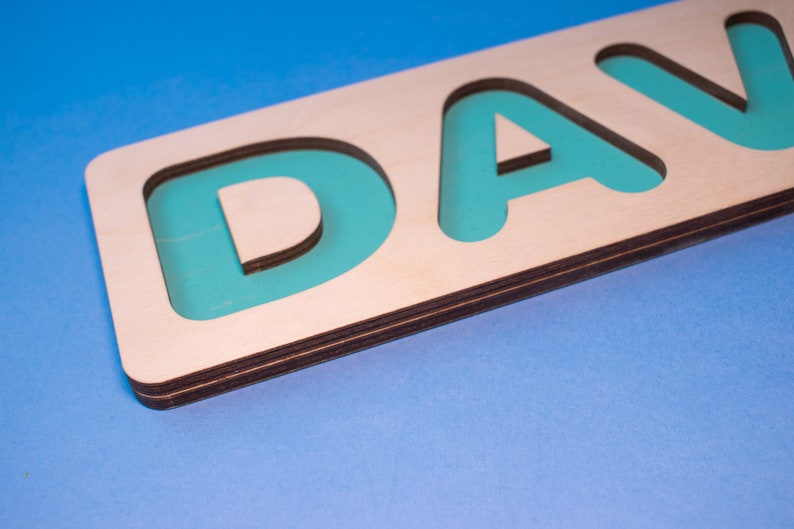 Op de afbeelding: Houten naam puzzel met de letters "DAVID" in turquoise in een natuurlijke houten lijst. De puzzel is ontworpen met een lichtblauwe achtergrond. De letters zijn uitgesneden, waardoor een verzonken gebied voor elke letter ontstaat.
