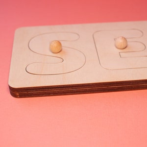 Op de afbeelding: Houten puzzel met de letters "SEE" in een lichte houtafwerking. De puzzelstukken hebben de vorm van de letters, met kleine houten knoppen voor een gemakkelijke bediening. De achtergrond is effen koraal van kleur.