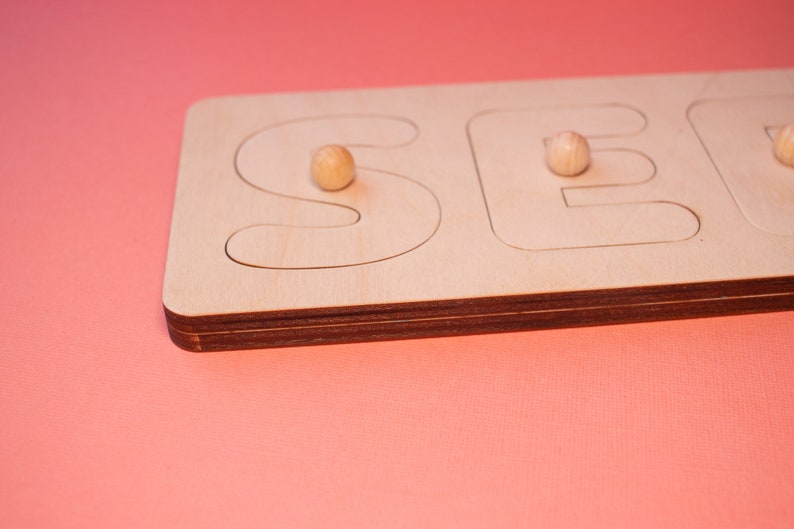 Op de afbeelding: Houten puzzel met de letters "SEE" in een lichte houtafwerking. De puzzelstukken hebben de vorm van de letters, met kleine houten knoppen voor een gemakkelijke bediening. De achtergrond is effen koraal van kleur.