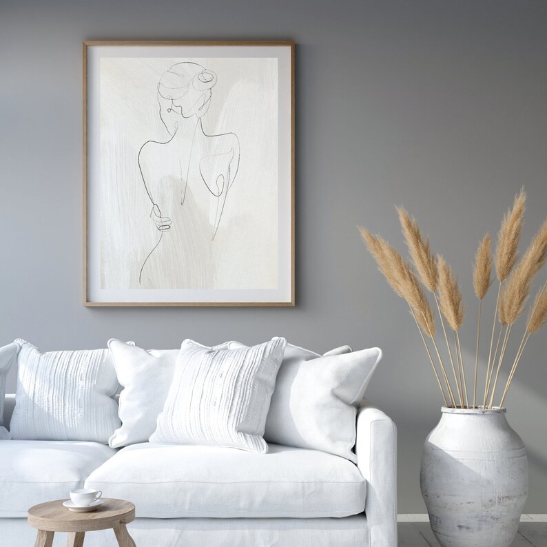 May include: A white couch with three white pillows in front of a gray wall. A framed black and white line drawing of a woman is hanging on the wall. A vase with dried pampas grass is on the floor next to the couch.