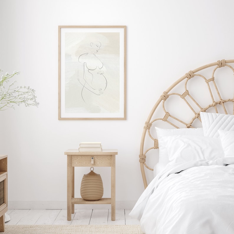 May include: A framed line drawing of a pregnant woman in a minimalist style, with a beige background. The frame is hanging on a white wall above a wooden nightstand with a woven basket. A white bed with a wicker headboard is in the background.