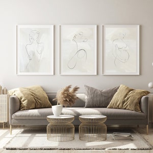 May include: Three framed line drawings of female figures in various poses on a white wall above a gray couch with pillows and two gold metal side tables. The drawings are on a beige background.