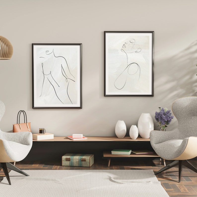 May include: Two framed black and white line drawings of women's figures on a beige wall. The drawings are in a minimalist style. The wall is above a long wooden shelf with a few decorative items, including a vase and a book. Two chairs are in front of the shelf, one with a brown leather bag on it.
