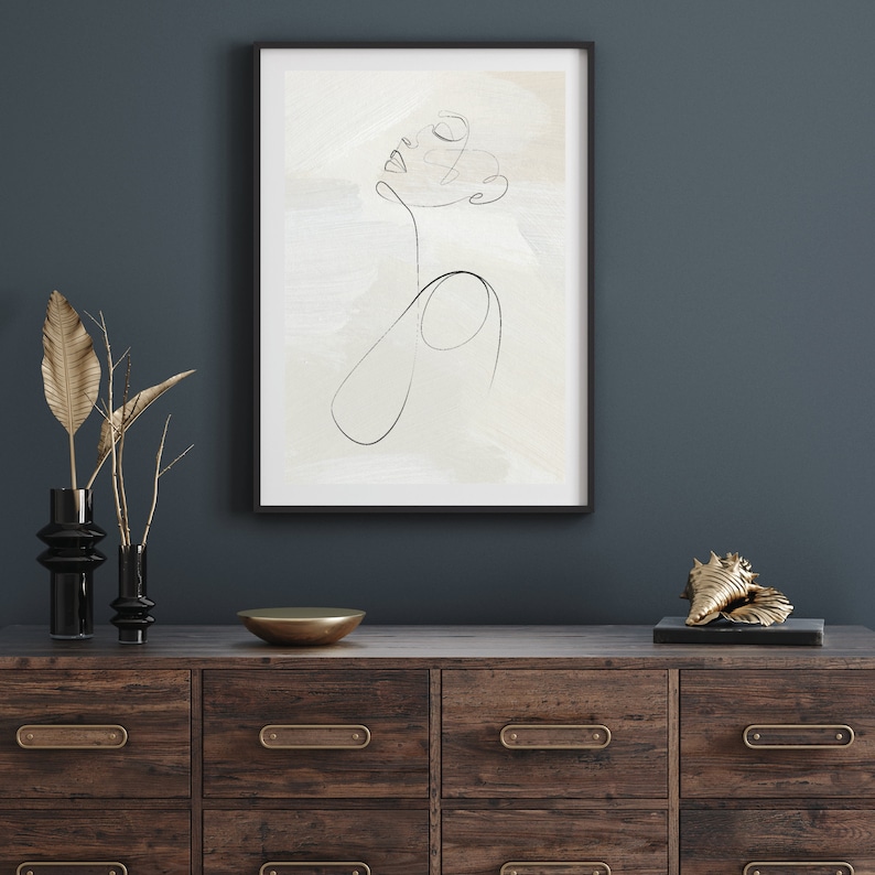 May include: A black framed print of a line drawing of a face on a white background with a light brown wash. The print is hanging on a dark blue wall above a wooden dresser with six drawers.