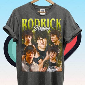 May include: Dark gray t-shirt featuring a collage of images and the text "RODRICK Heffley" in yellow. The shirt has a vintage style and is made of a soft material. The shirt is hanging on a wooden hanger.