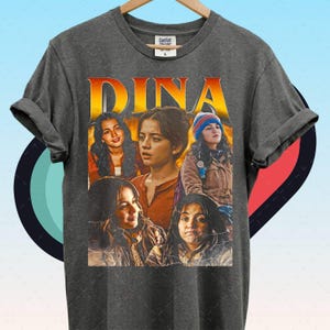 May include: Charcoal grey t-shirt with a collage of images of a person, and the name "DINA" in orange. The images have a vintage, distressed appearance. The t-shirt has a crew neck and short sleeves.