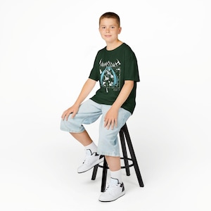 May include: Dark green t-shirt featuring a graphic of a female anime character with blue hair and the text "Lingua Ignota". The shirt is worn with light blue denim shorts and white sneakers. The person is sitting on a black stool.