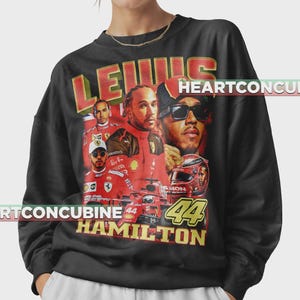 Pode incluir: Camisola preta com uma colagem de imagens de Lewis Hamilton, piloto de F&oacute;rmula 1. O design inclui o nome "LEWIS" em letras vermelhas e douradas, juntamente com "HAMILTON" e o n&uacute;mero "44". As imagens mostram Hamilton em trajes de corrida.