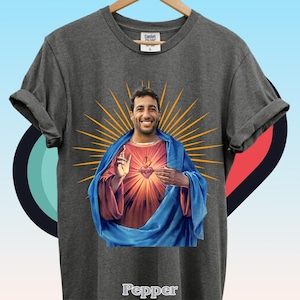 May include: A charcoal gray short-sleeve t-shirt featuring a graphic print of a smiling man with a beard, depicted in the style of a religious icon with a blue robe, red tunic, and a radiant orange sunburst background. The word Pepper is printed in white.