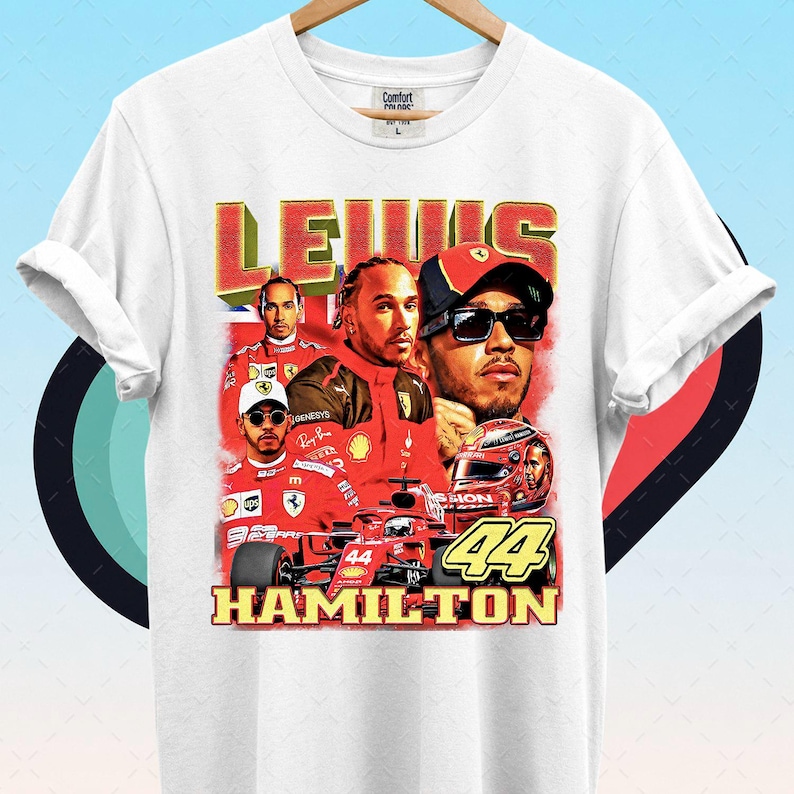 Pode incluir: Camiseta branca com um gr&aacute;fico de Lewis Hamilton em traje de corrida vermelho, com o nome "LEWIS" em vermelho e amarelo, e "HAMILTON" abaixo. O n&uacute;mero "44" &eacute; exibido de forma proeminente.