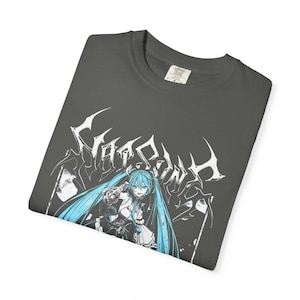 May include: Dark gray t-shirt featuring a graphic of an anime character with long, light blue hair. The word "CYBERPUNK" is printed above the character in a stylized font. The shirt is folded and has a tag in the upper right corner.