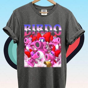 May include: Dark grey t-shirt with a graphic of the character Birdo from the Super Mario series. The design features the word "BIRDO" in a gradient font, and multiple pink Birdo characters with red bows.