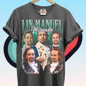 Puede incluir: Camiseta gris oscuro con un collage de imágenes de Lin-Manuel Miranda y el texto "LIN-MANUEL Miranda" en un diseño verde y blanco. La camiseta está colgada en una percha de madera.