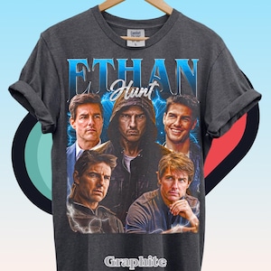 Może przedstawiać: Ciemnoszary t-shirt z kr&oacute;tkim rękawem z nadrukiem graficznym przedstawiającym Ethana Hunta. Wz&oacute;r zawiera pięć r&oacute;żnych wizerunk&oacute;w postaci, napis "ETHAN" dużymi niebieskimi literami, napis "Hunt" oraz błyskawice na ciemnym tle.