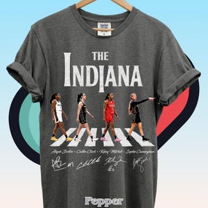 May include: A charcoal gray t-shirt featuring the text THE INDIANA in white, with a graphic of four basketball players walking across a crosswalk. Below the players are the names Aliyah Boston, Caitlin Clark, Kelsey Mitchell, and Sophie Cunningham, along with their respective signatures and jersey numbers. The shirt has a crew neck and short sleeves.