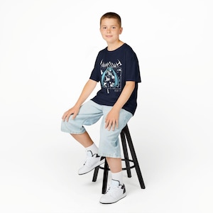May include: A navy blue t-shirt featuring a graphic design with a blue-haired anime character and Japanese text. The person is wearing light blue denim shorts and white sneakers with black accents. They are sitting on a black stool against a white background.