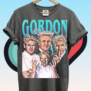 May include: Dark gray t-shirt featuring a graphic of Gordon Ramsay with the text "GORDON Ramsay" in a retro font. The shirt displays multiple images of the celebrity chef. The shirt is hanging on a wooden hanger.