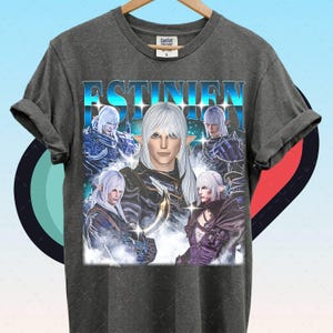 Puede incluir: Camiseta gris oscuro con un gráfico de un personaje con cabello blanco y el nombre "ESTINIEN" en azul. El diseño incluye múltiples imágenes del personaje en varias poses. La camiseta está colgada en una percha de madera.