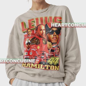 Pode incluir: Camisola bege com um gr&aacute;fico de Lewis Hamilton, com o texto "LEWIS HAMILTON 44" em vermelho e amarelo. O design inclui imagens de um carro de corrida e um capacete.