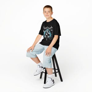 May include: Black t-shirt featuring a graphic design with a blue-haired anime character and Japanese text. The shirt is worn with light blue denim shorts and white sneakers. The person is sitting on a black stool against a white background.