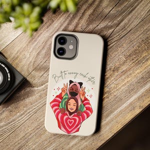 以下が含まれることがあります： ベージュ色の携帯電話ケース。ヒジャーブとセーターを着た3人の人物がピースサインをしているイラストが描かれています。「But to annoy each other」というテキストが筆記体で書かれています。ケースにはカメラ用の切り抜きがあります。木製の表面にあります。