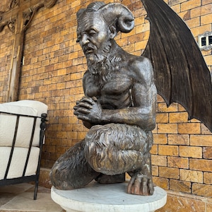 May include: Bronze statue of a kneeling satyr with horns, wings, and a beard, atop a white marble pedestal. The statue is set against a brick wall with a wooden cross in the background. The satyr's hands are clasped in prayer.