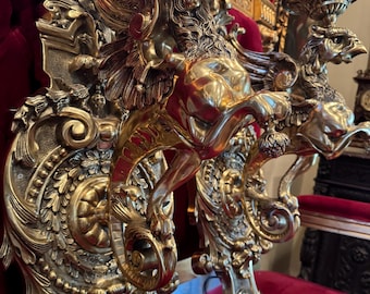 May include: Ornate, gold-colored wall sconces with detailed griffin and floral designs. The sconces feature a candle holder and are mounted on a red velvet surface. The design is reminiscent of antique decor.
