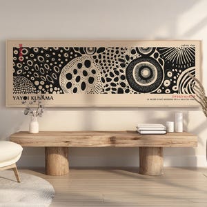 Könnte beinhalten: Ein gerahmter Kunstdruck mit einem schwarz-weißen abstrakten Design, das kreisförmige und gepunktete Muster aufweist. Der Druck enthält den Text "YAYOI KUSAMA" und "JAPAN 1986". Das Kunstwerk wird über einer Holzbank ausgestellt.
