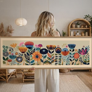 May include: A framed floral art print featuring a colorful array of stylized flowers in shades of orange, blue, pink, and green. The artwork has a beige background and a light brown wooden frame. The print is held by a person.