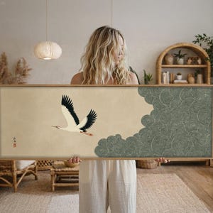May include: A framed artwork featuring a crane in flight against a beige background. The crane has black and white wings and a red beak. A textured, gray cloud design is on the right side. The frame is made of wood.