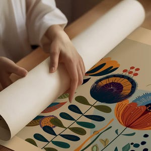 May include: A rolled-up art print with a floral design is being unrolled. The print features large, stylized flowers in shades of orange, blue, and purple, with green leaves and stems against a light background. The print is being unrolled on a wooden surface.