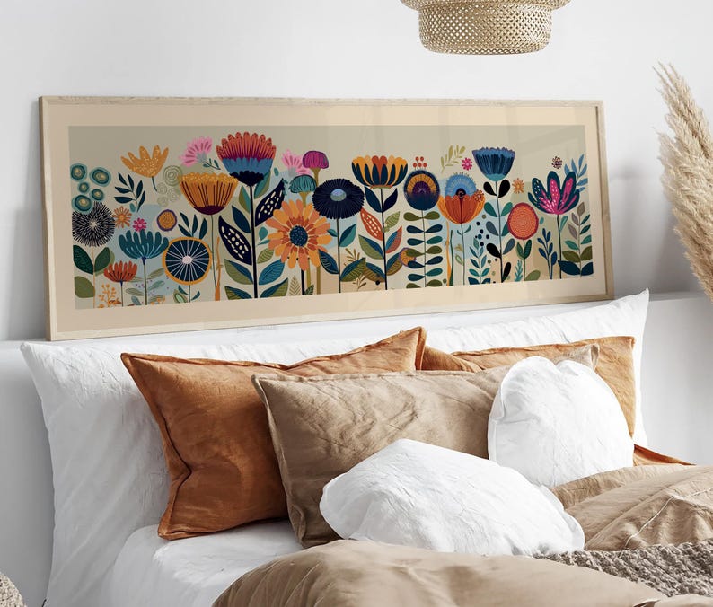 May include: A framed floral art print featuring a colorful array of stylized flowers in shades of blue, orange, pink, and yellow. The artwork is set against a light beige background and framed in a light wood tone. The print is displayed above a bed with pillows.
