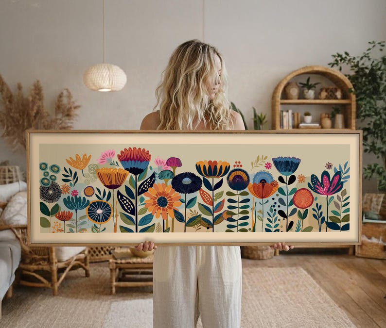 May include: A framed floral art print featuring a colorful array of stylized flowers in shades of orange, blue, pink, and green. The artwork has a beige background and a light brown wooden frame. The print is held by a person.