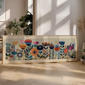 May include: A framed floral art print featuring a colorful array of stylized flowers in shades of blue, orange, pink, and green. The artwork is set against a light background and framed in a gold-colored frame. The print is displayed on a wooden floor.