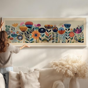 May include: A long, rectangular framed art print featuring a colorful floral design. The artwork showcases stylized flowers in shades of blue, orange, yellow, and pink against a light background. The frame is white. The artwork is being hung on a wall.
