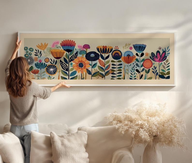 May include: A long, rectangular framed art print featuring a colorful floral design. The artwork showcases stylized flowers in shades of blue, orange, yellow, and pink against a light background. The frame is white. The artwork is being hung on a wall.