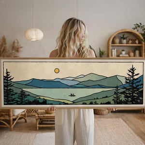 May include: A framed landscape art print featuring a serene mountain and lake scene. The artwork showcases a color palette of blues, greens, and a yellow sun, with dark green trees framing the view. The print is in a natural wood frame.