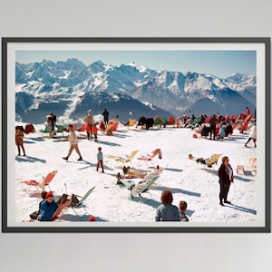 May include: A framed photographic print depicting a snowy mountain landscape with numerous people relaxing in colorful folding chairs. The scene features bright white snow, clear blue skies, and distant jagged mountain peaks under sunlight.