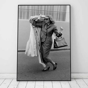 May include: A black and white photograph of a woman wearing a suit and sunglasses, carrying a large white garment and a black leather handbag. She is walking down a street.