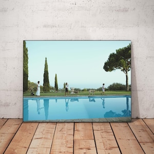 Könnte beinhalten: Ein gerahmtes Landschaftsfoto, das einen rechteckigen Swimmingpool mit klarem, blauem Wasser zeigt, in dem sich der Himmel spiegelt. Neben dem Pool gehen drei Personen und zwei Hunde auf einem Grasweg, der von Zypressen und einer Kiefer gesäumt ist.