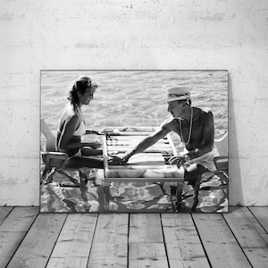 May include: Black and white photograph of a framed image depicting two people playing backgammon in shallow water. The man wears a hat and necklace, while the woman is in a bikini. The game board is in the center.
