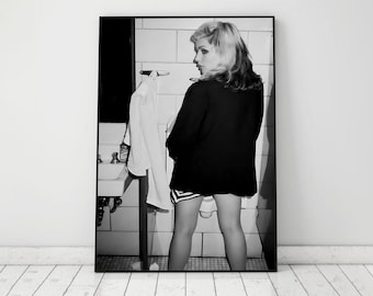 May include: A black and white photo of a person standing in a bathroom. The person is wearing a black jacket and a skirt. The person is looking over their shoulder at the camera.