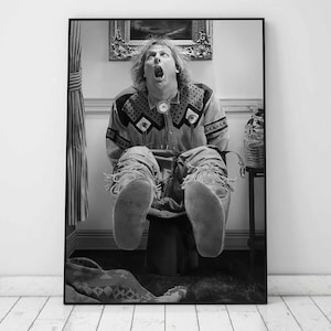 Pode incluir: Uma fotografia a preto e branco de uma pessoa sentada num vaso sanitário com as pernas levantadas no ar. A pessoa está a usar uma camisa e calças estampadas. A fotografia está enquadrada numa moldura preta.