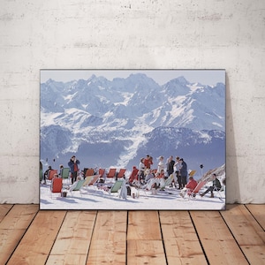 May include: A framed print featuring a vintage photograph of a snowy mountain scene. People are relaxing in colorful lounge chairs on a snow-covered slope. The background showcases majestic snow-capped mountains under a clear sky.