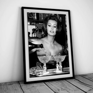 May include: Black and white photo of a woman pouring champagne into two glasses. The woman is wearing a low-cut dress and has her hair styled in a bouffant. The photo is framed in a black frame.