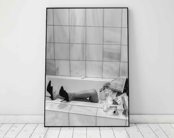 May include: Black and white photograph of a person in a white bathrobe relaxing in a bathtub. The person is holding a newspaper in front of their face and has their feet resting on the edge of the tub. The bathtub is in a tiled bathroom.
